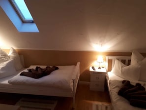 Une petite pièce à la lumière tamisée, avec deux lits, une lucarne et une table de chevet sur laquelle se trouve une lampe.