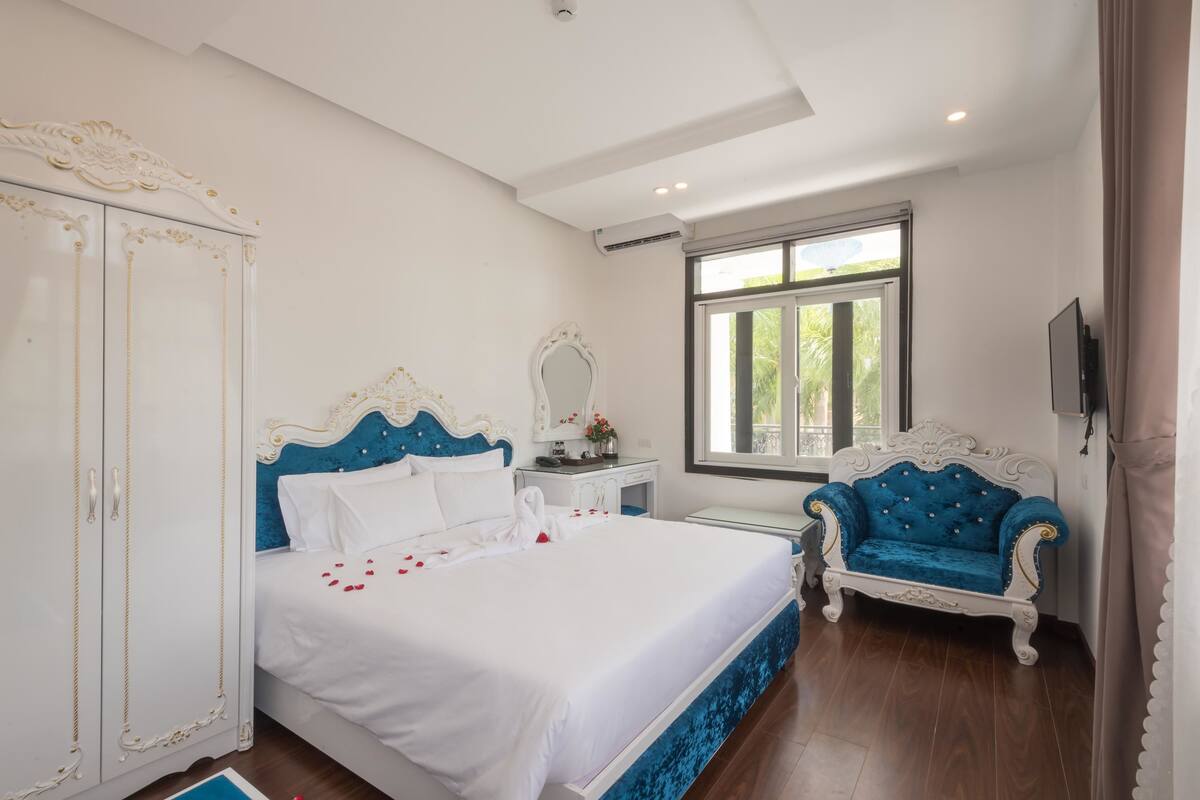 A bedroom with a large bed, a wardrobe, a vanity with a mirror, a blue armchair, and a window with a view of greenery.