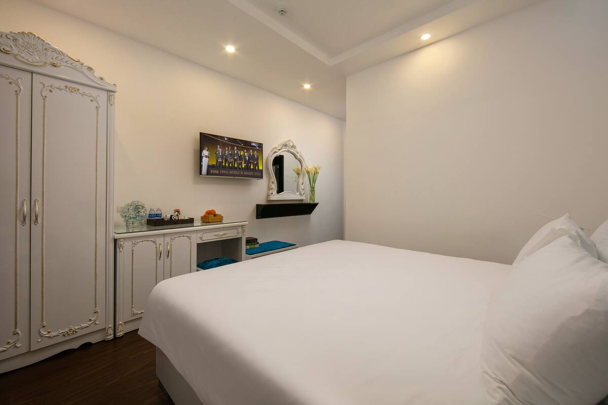 A bedroom with a large bed, a wardrobe, a vanity with a mirror, and a shelf with decorative items.