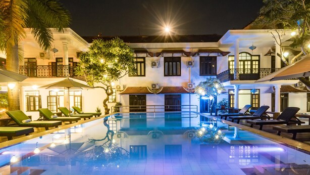 A swimming pool with lounge chairs and umbrellas, surrounded by buildings with balconies and windows. - Hoian Nostalgia Hotel & Spa (Da Nang)