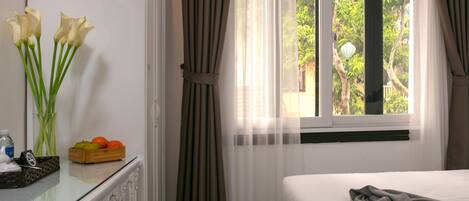 A hotel room with a bed, a desk with a vase of flowers, a fruit bowl, and a window with curtains.