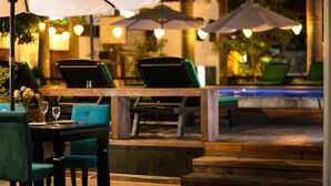 An outdoor seating area with wooden tables and chairs, umbrellas, and a pool in the background. - Hoian Nostalgia Hotel & Spa (Da Nang)