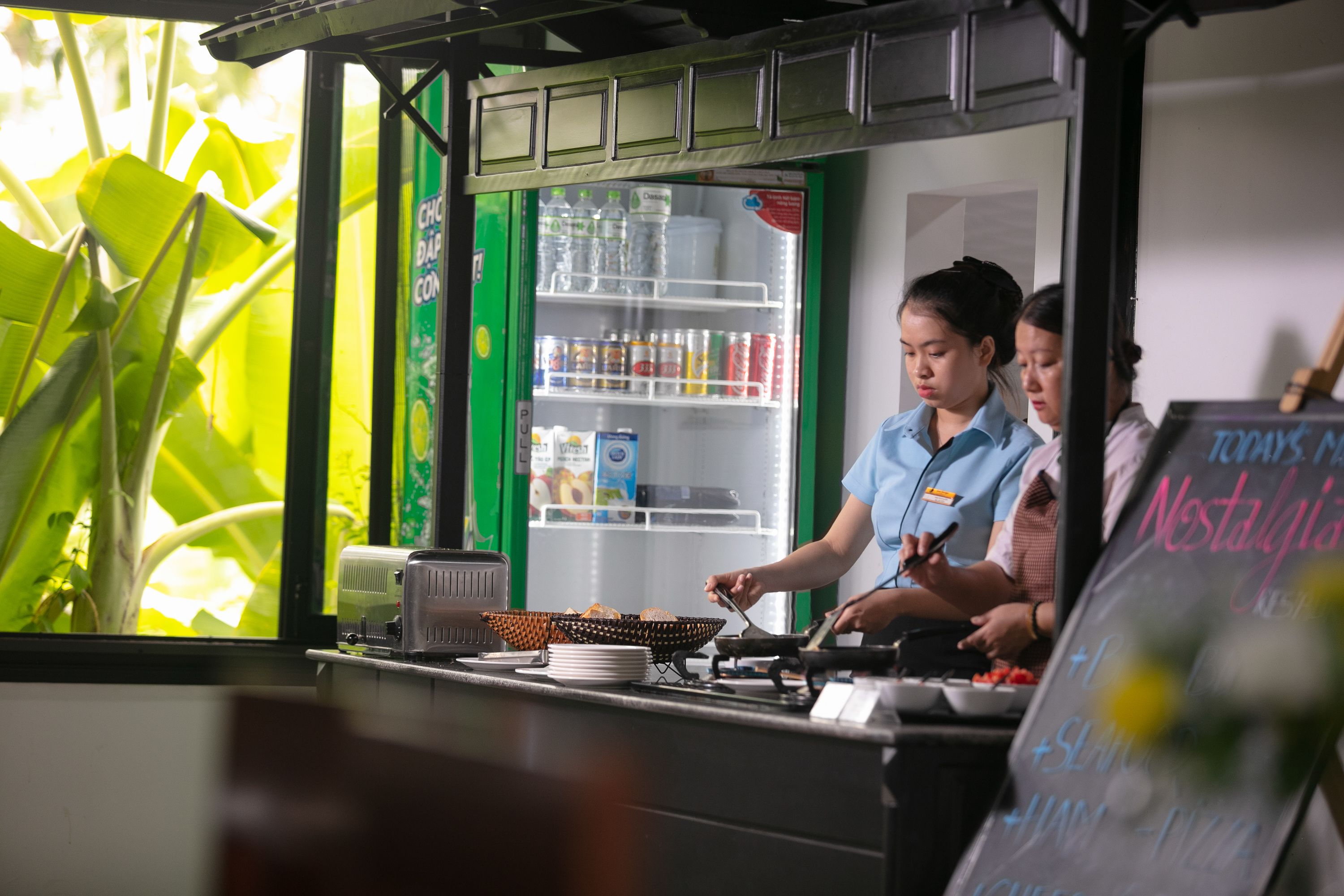 A food stall with a refrigerator stocked with beverages and snacks, a counter with bowls of food, and a menu board.