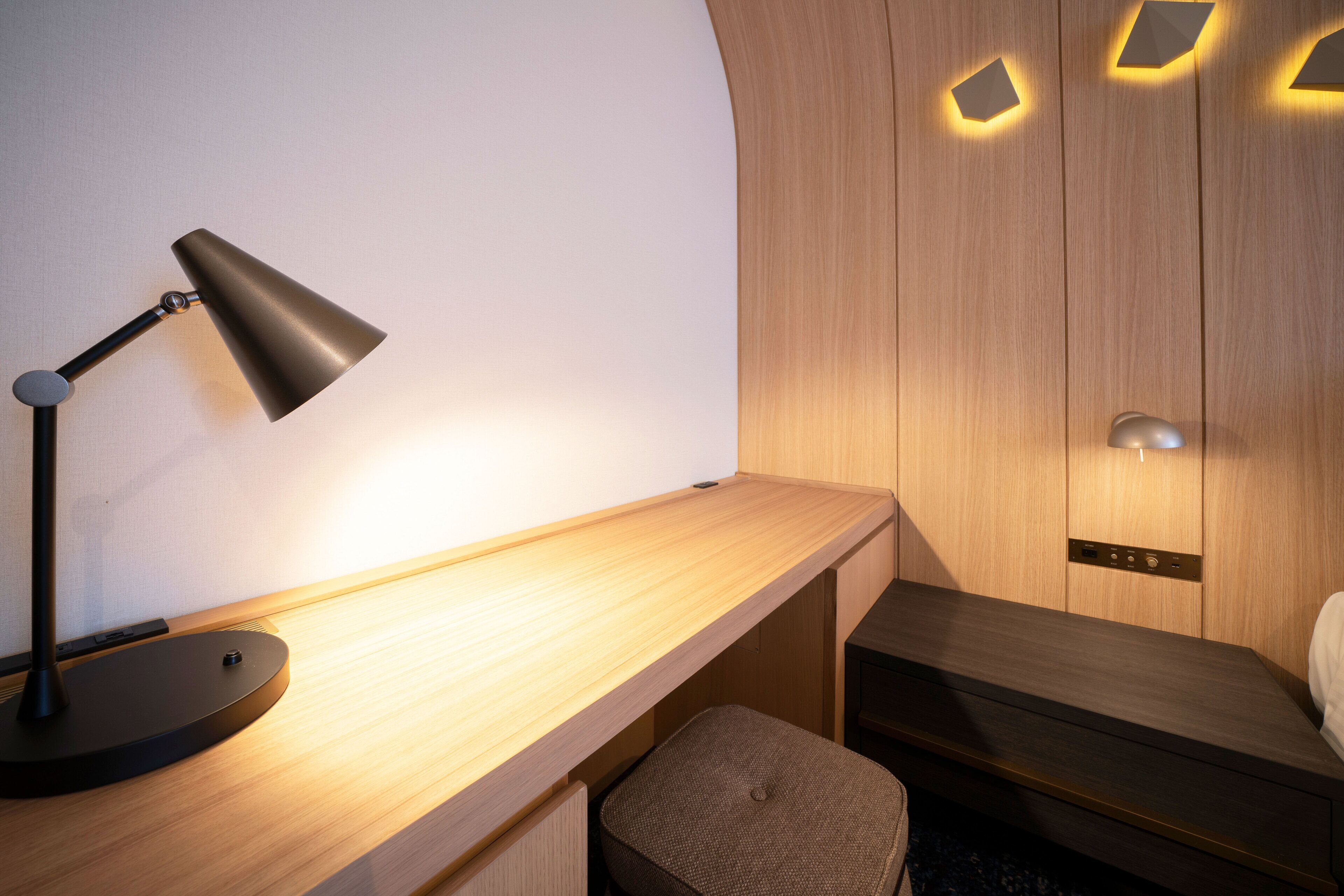 Een moderne kamer met een houten bureau, een zwarte bureaulamp, een klein gestoffeerd krukje en een bed met een houten hoofdeinde.