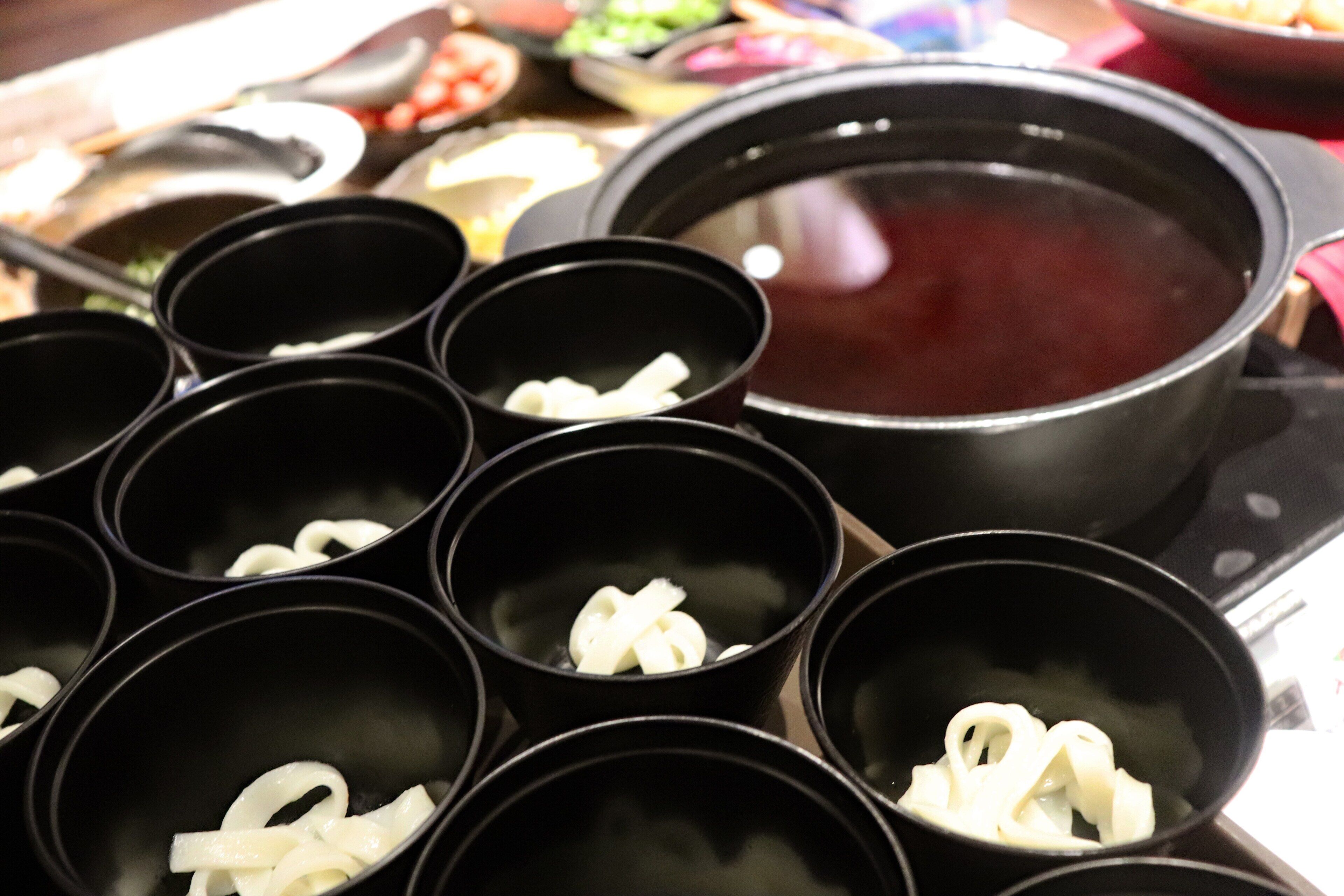 A table with several black bowls containing white noodles, a large black pot with a red liquid, and various other dishes in the background.