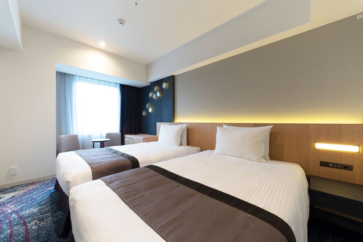 Une chambre d’hôtel avec deux lits, un bureau, une chaise, une petite table et une fenêtre avec des rideaux.
