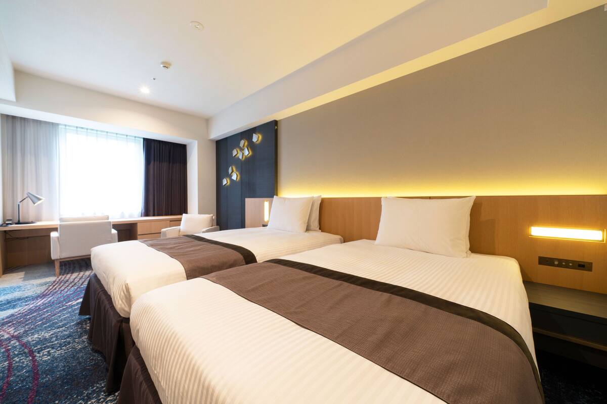 Une chambre d’hôtel avec deux lits, un bureau, une chaise et une grande fenêtre avec des rideaux.
