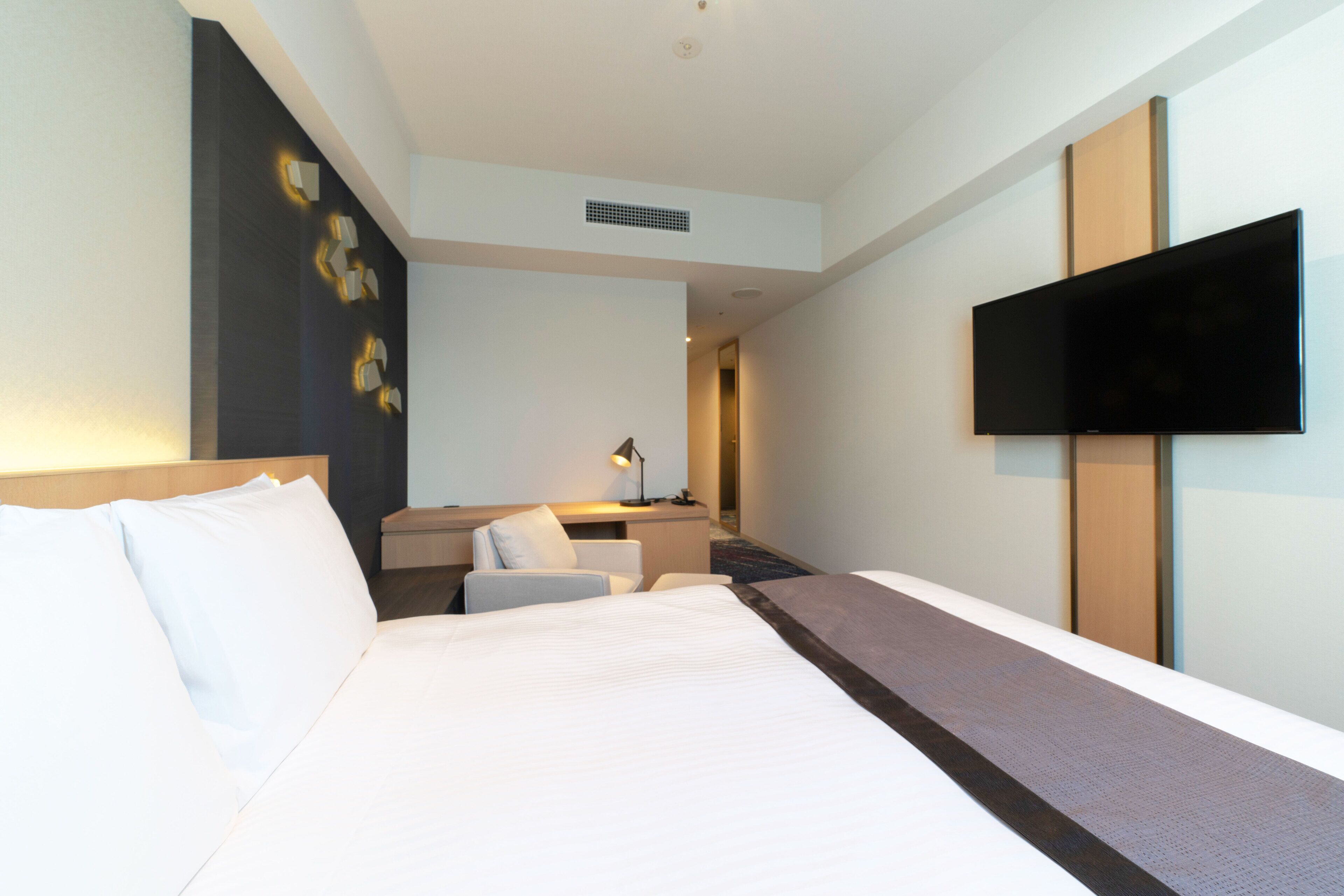 A hotel room with a large bed, a desk with a chair, a flat-screen TV, and a wall-mounted TV.