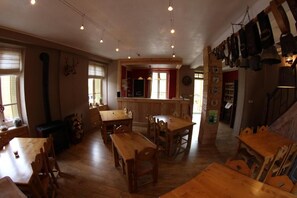 A spacious dining area with wooden tables and chairs, a bar area with stools, and a kitchen visible in the background. - Le silence du midi (Comus)