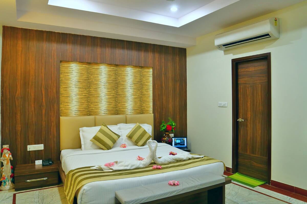 A hotel room with a bed, bedside table, nightstand, air conditioning unit, and a wooden door.