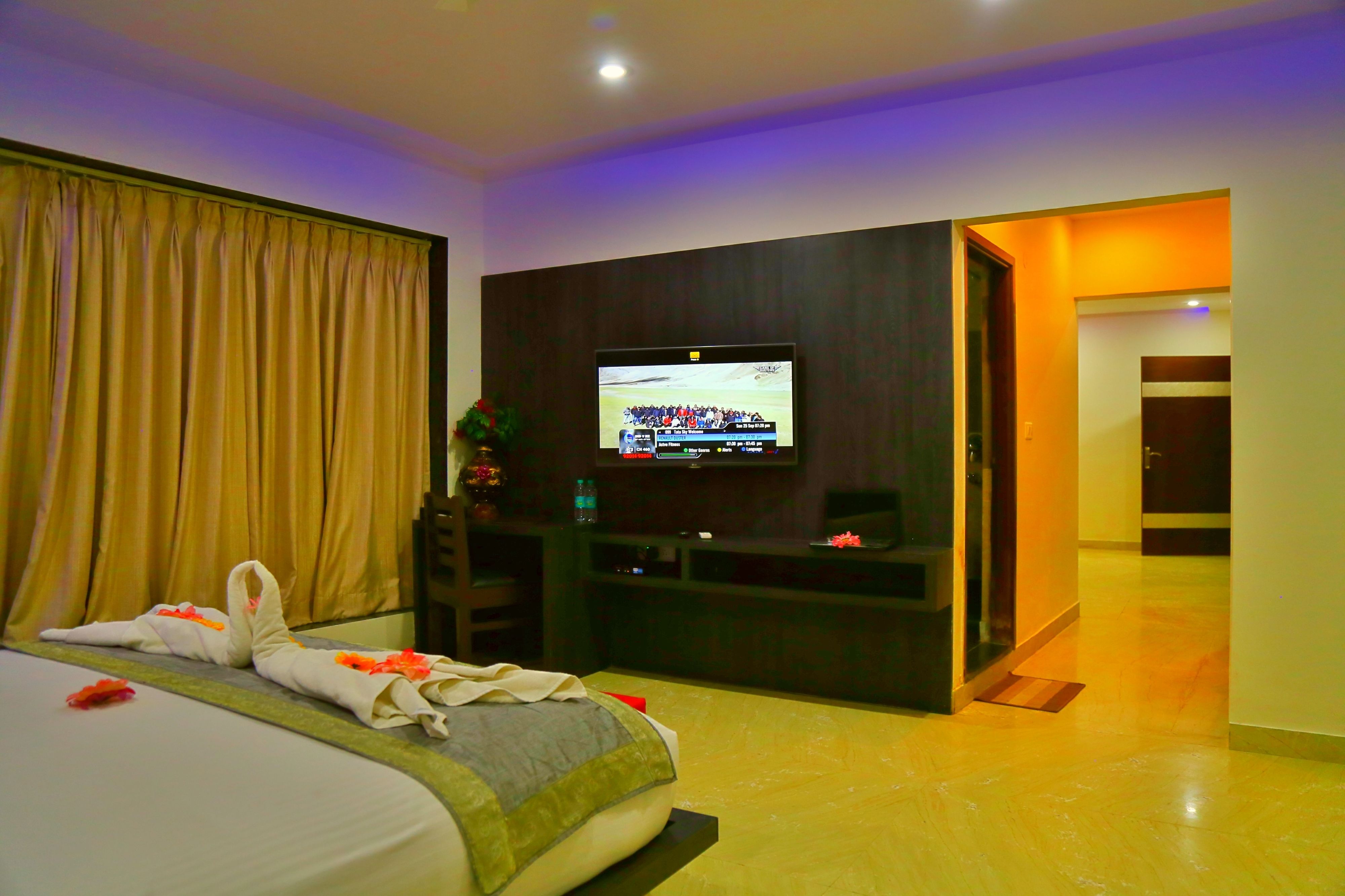 A hotel room with a bed, a flat-screen TV mounted on the wall, a desk, and a door leading to another room.