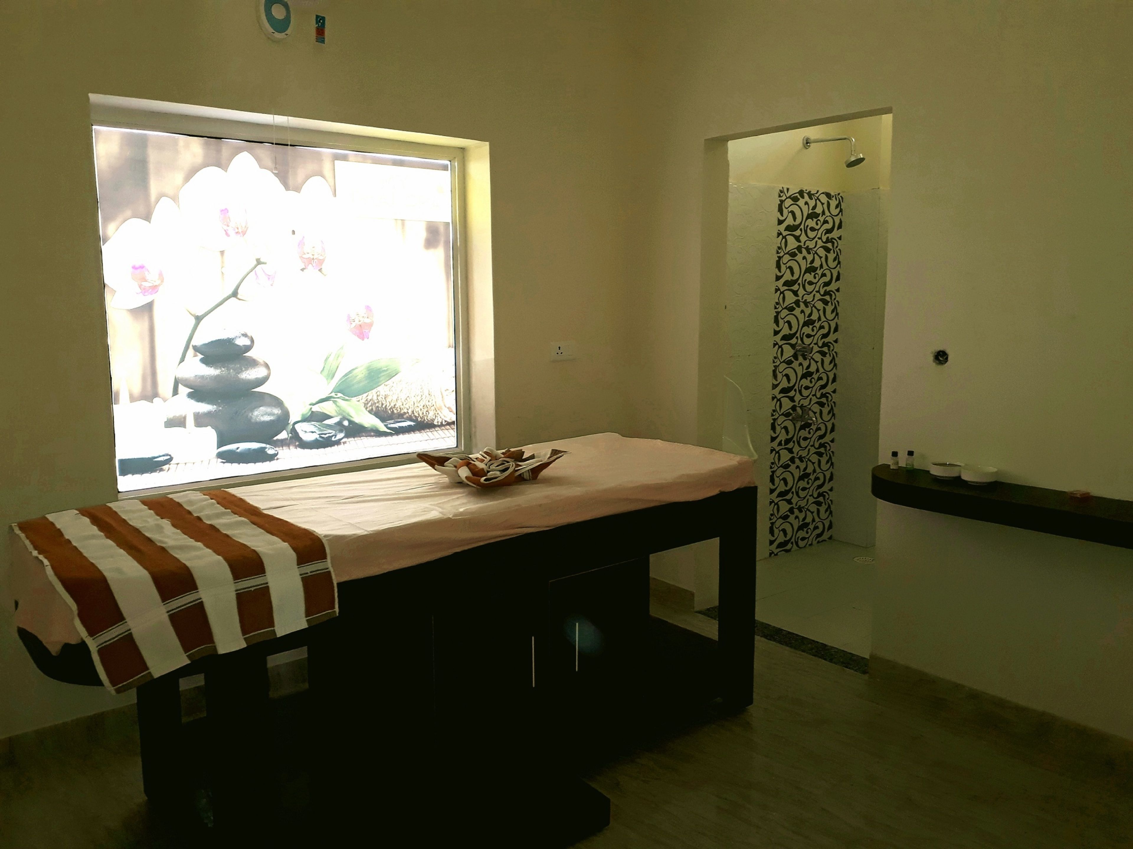 A room with a bed, a small table with a bowl, a window with a view of rocks and flowers, and a shower area with a patterned curtain.