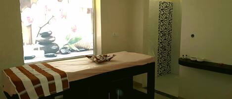 A room with a bed, a small table with a bowl, a window with a view of rocks and flowers, and a shower area with a patterned curtain.