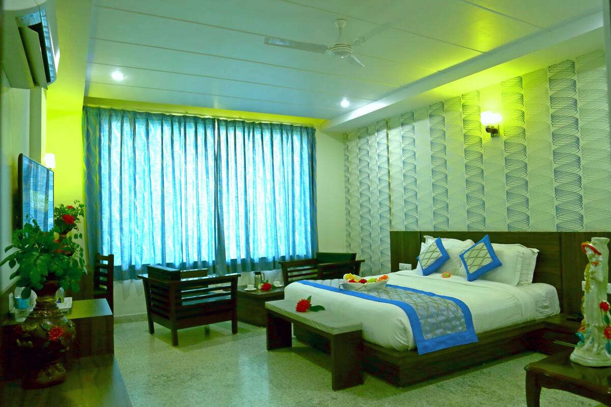 A hotel room with a bed, bedside tables, a desk, a chair, a potted plant, and a large window with curtains.