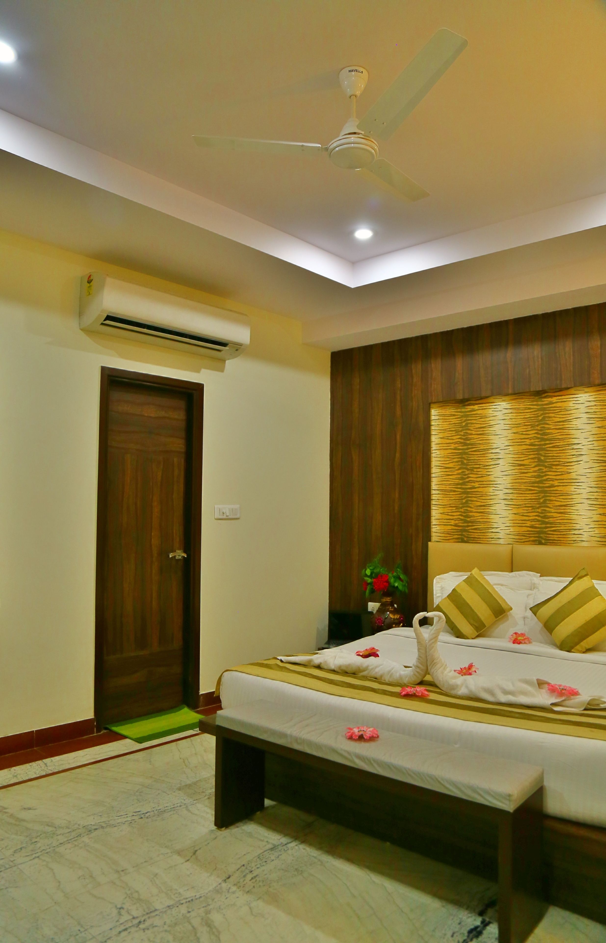 Una habitación de hotel con cama, ventilador de techo, equipo de aire acondicionado, puerta de madera y un panel decorativo en la pared.
