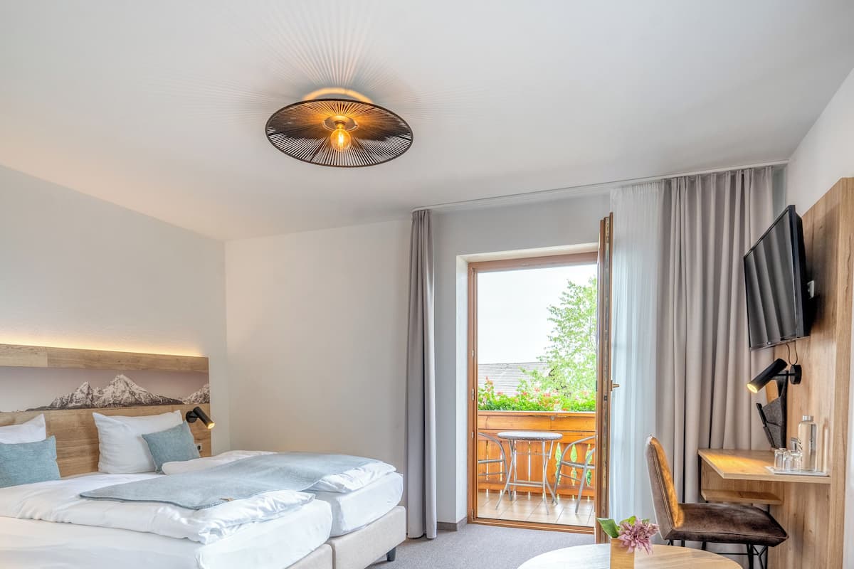 A hotel room with a bed, a desk with a chair, a TV, and a balcony with a view of greenery.