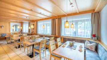 A wooden-paneled dining area with tables and chairs, large windows with curtains, and hanging pendant lights.