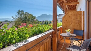 Een houten balkon met uitzicht op bergen, een tafeltje met een plant en rieten stoelen.