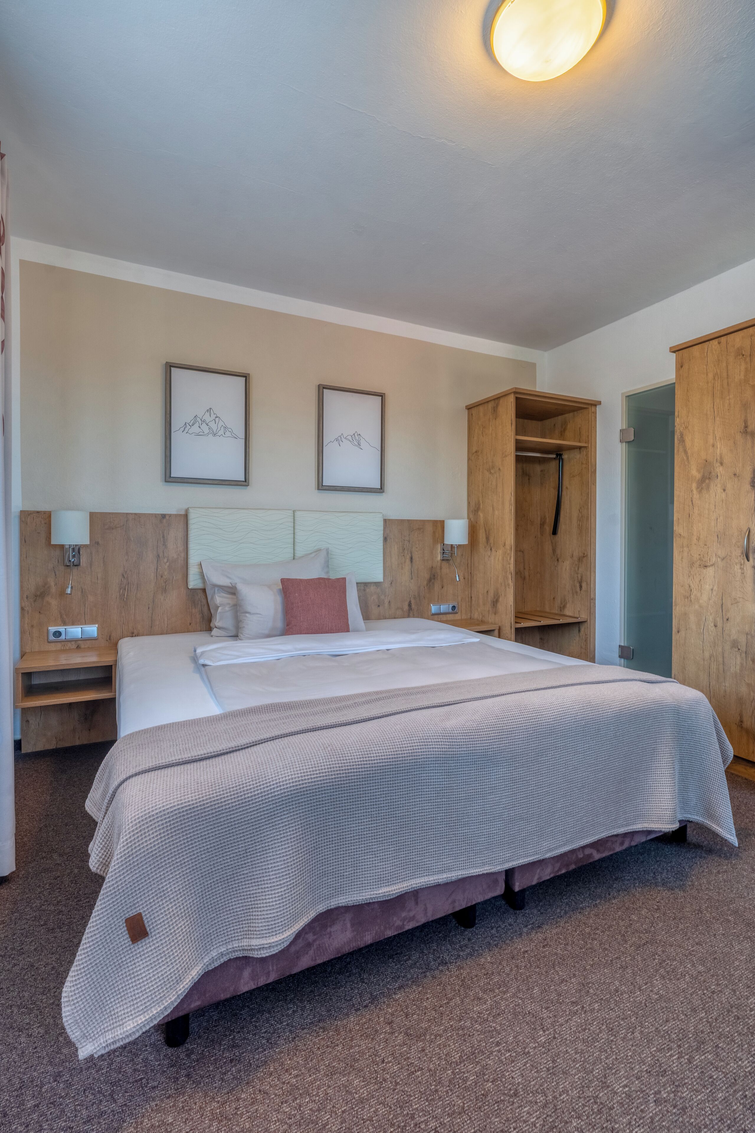 A hotel room with a large bed, wooden wardrobe, bedside tables, and wall-mounted artwork.