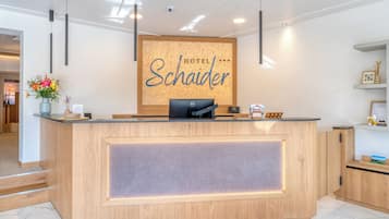 Een hotel receptiebalie met een bordje met de tekst 'HOTEL Schaider', een computer, een vaas met bloemen en een receptiegedeelte met een houten toonbank en kasten.