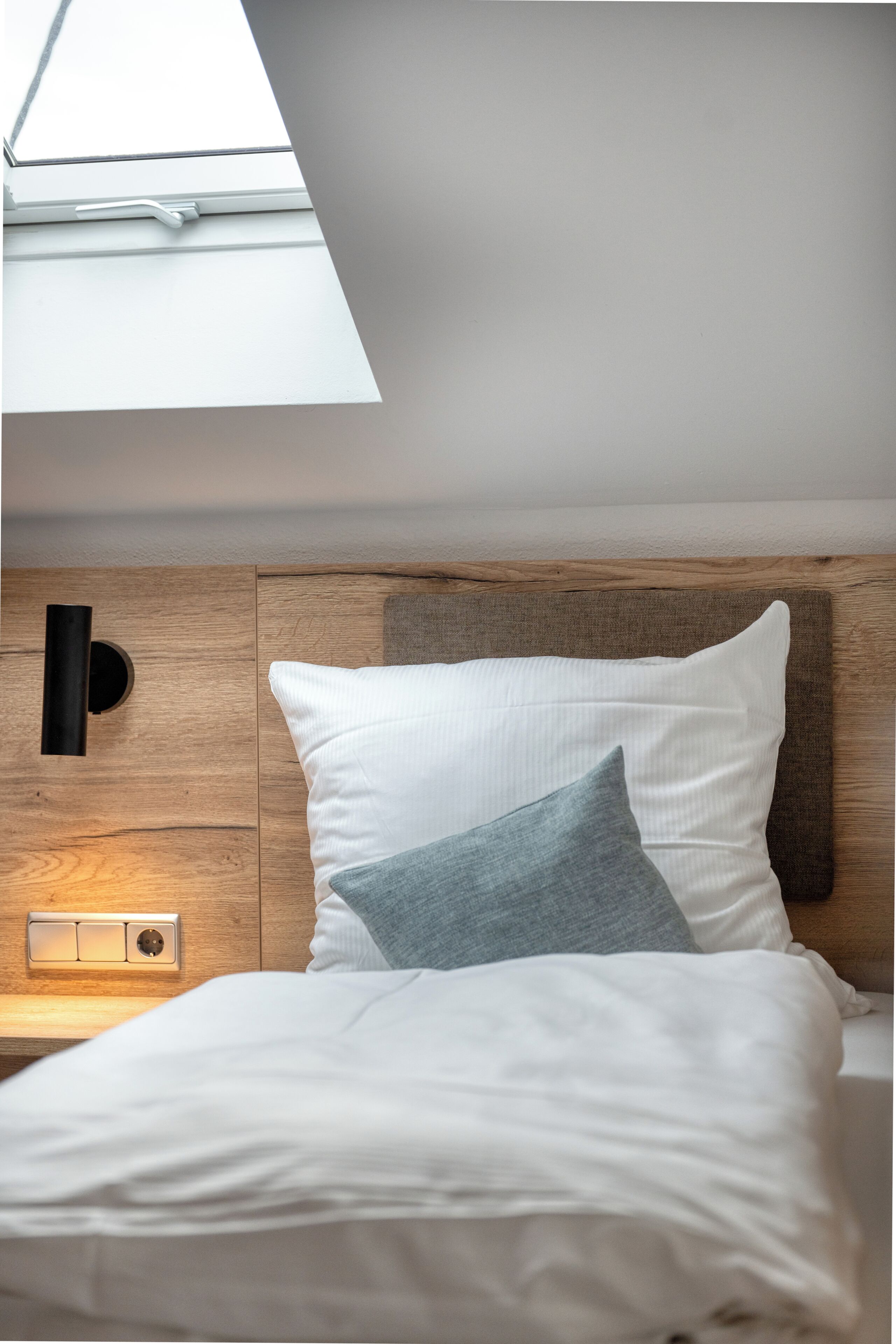 A bedroom with a skylight, a wooden headboard, a bedside table with a light, and a bed with white and grey bedding.