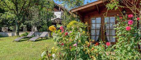 A wooden cabin with a garden in front, a lawn, and a tree.