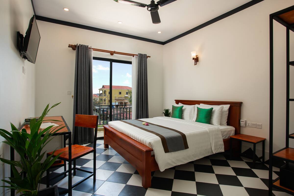 A hotel room with a checkered floor, a bed with a wooden headboard, a desk with a chair, a potted plant, and a balcony with curtains.