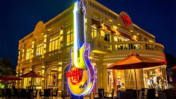 Ein Gebäude mit einem großen, gitarrenförmigen Neonzeichen, Außenplätzen und einem sichtbaren Restaurantbereich.