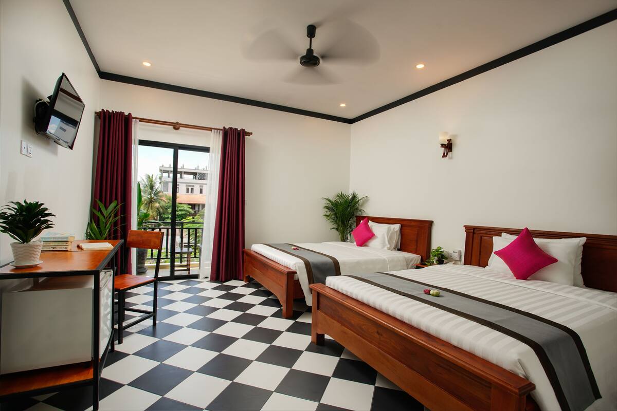 A hotel room with two beds, a desk, a chair, a TV, and a balcony with red curtains.