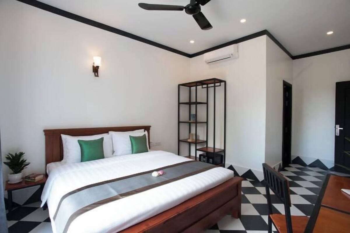 A hotel room with a bed, a ceiling fan, a small table, a chair, a shelf unit, and a checkered floor.