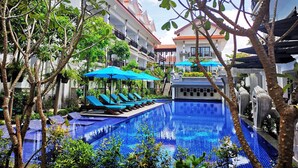Una piscina con baldosas azules, rodeada de árboles y sillones de jardín con sombrillas azules.