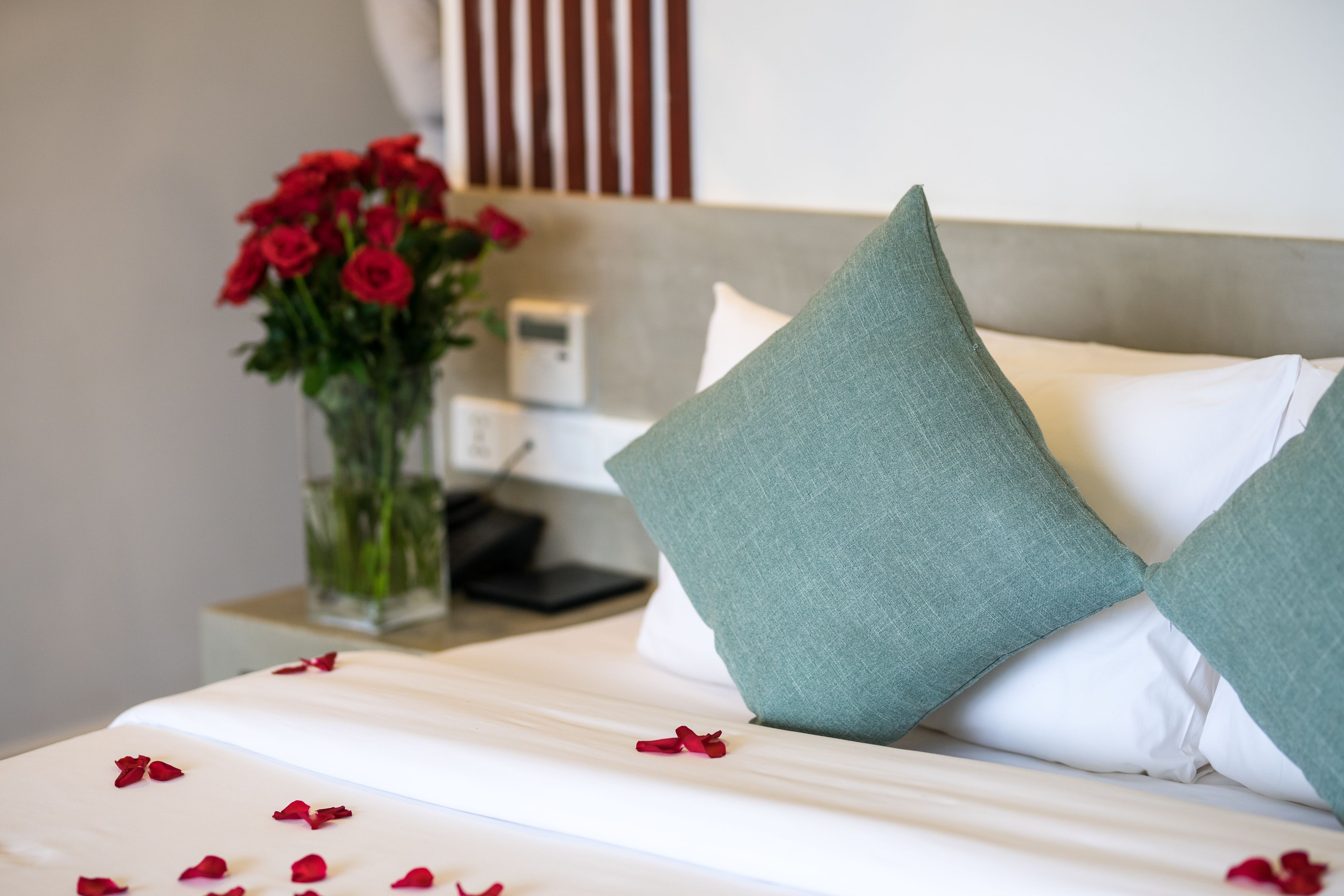 A neatly made bed with white linens and green pillows, a vase with red roses, and a bedside table with a lamp and a phone.