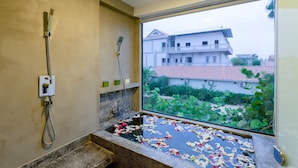 Un baño con ducha, lavamanos y ventana con vista a edificios y vegetación.