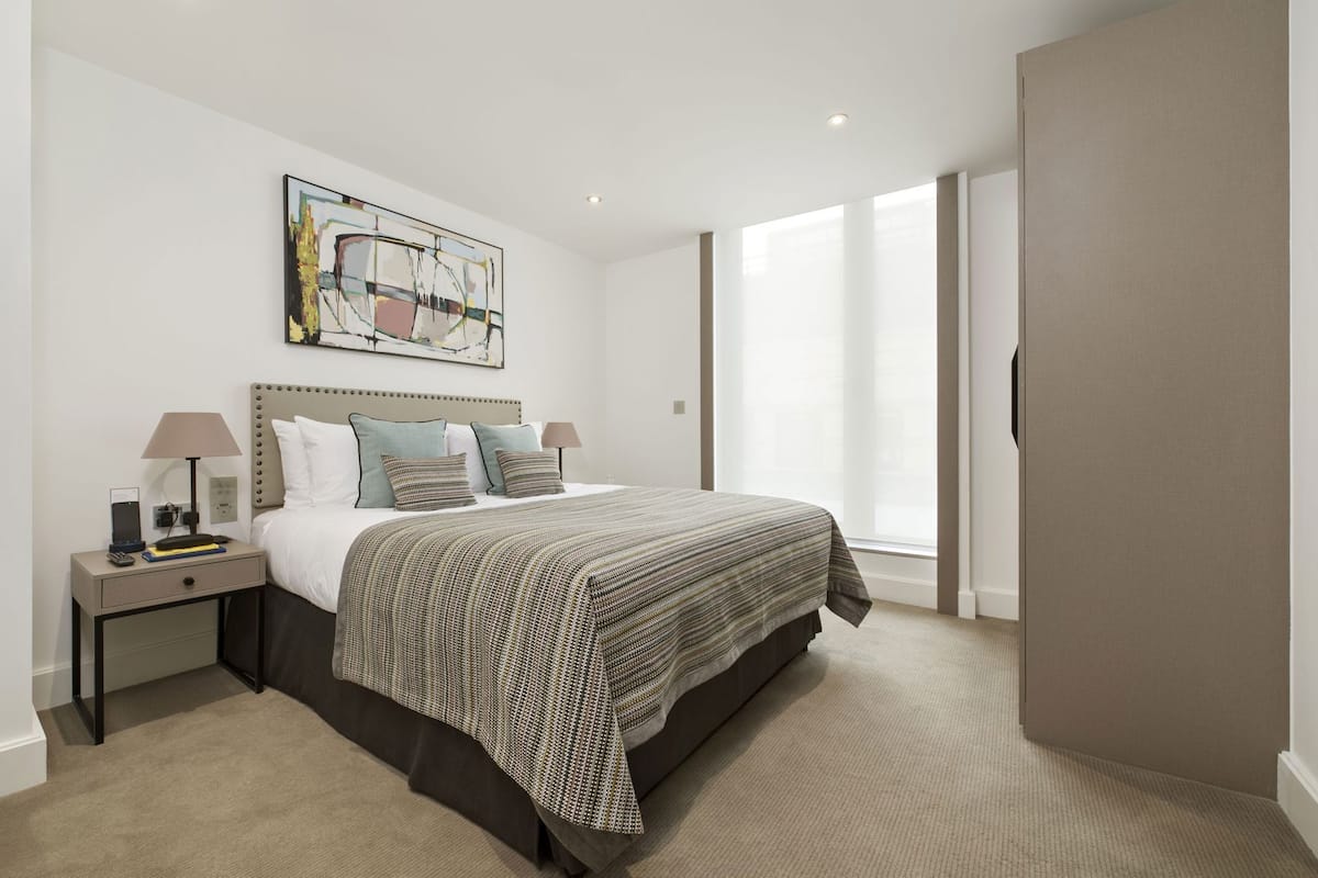 Habitación de hotel con una cama, una mesita de noche con lámpara, un cuadro en la pared y un ventanal grande.
