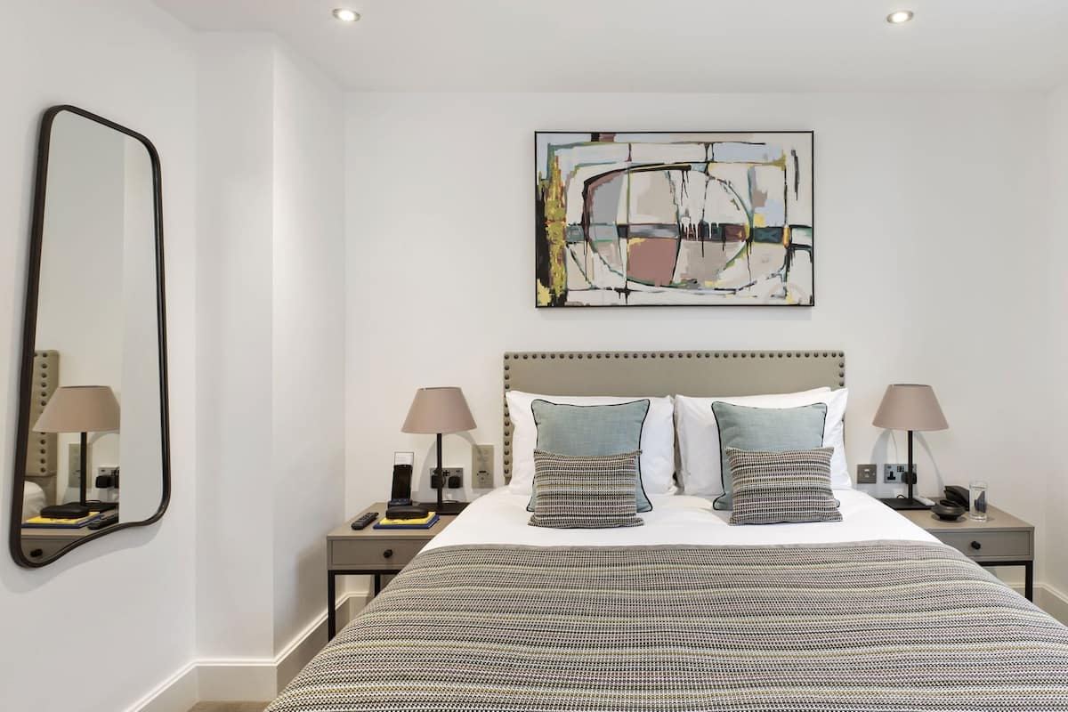 Una habitación de hotel con una cama grande, dos mesitas de noche con lámparas, un cuadro abstracto en la pared y un espejo amplio.