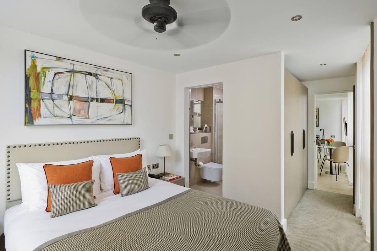 Una habitación de hotel con una cama grande, un ventilador de techo, un baño visible a través de una puerta abierta y un área de comedor al fondo.
