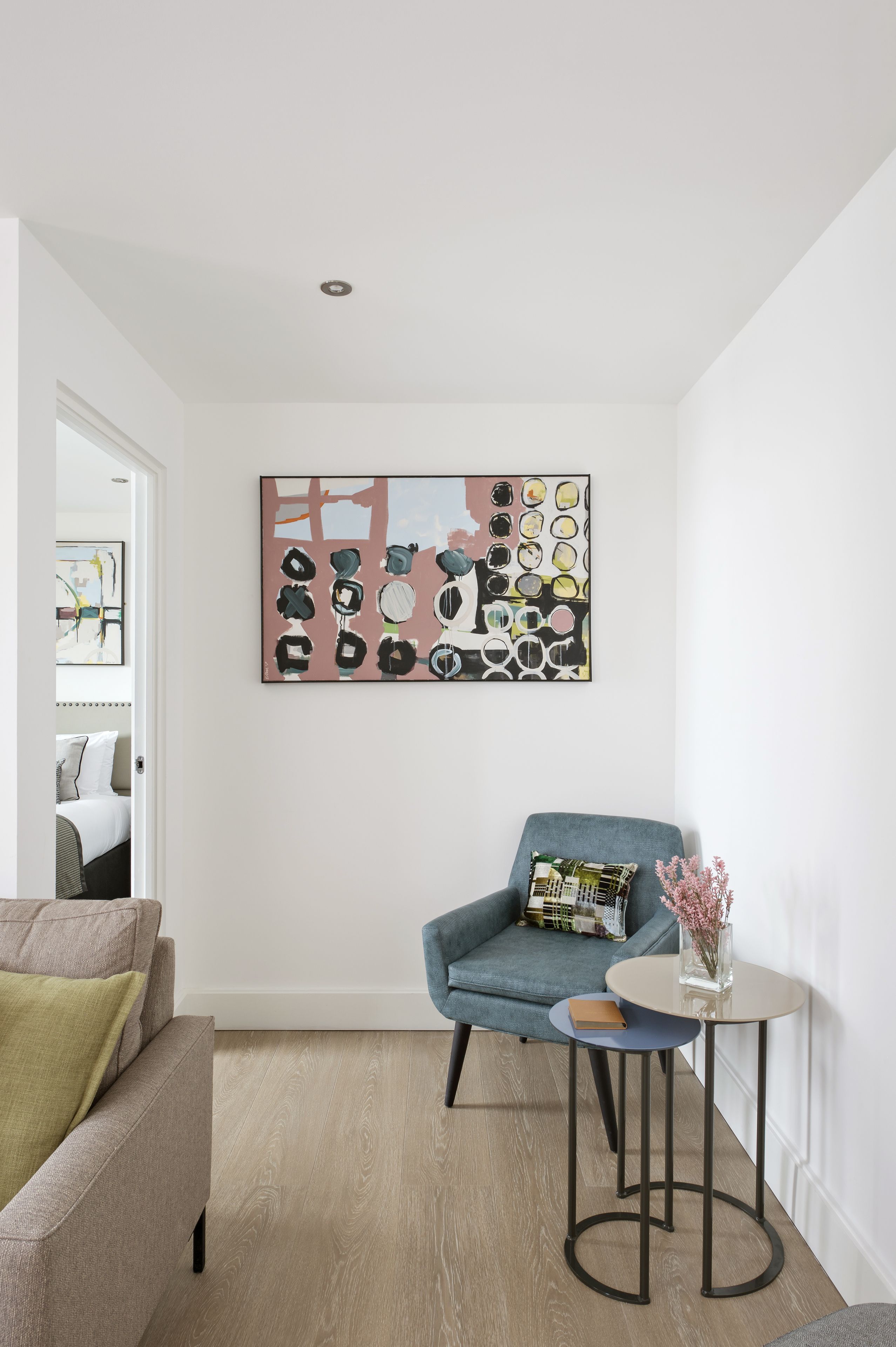 Een moderne woonkamer met een blauwe fauteuil, een klein rond tafeltje met een vaas, en een kleurrijk abstract schilderij aan de muur.