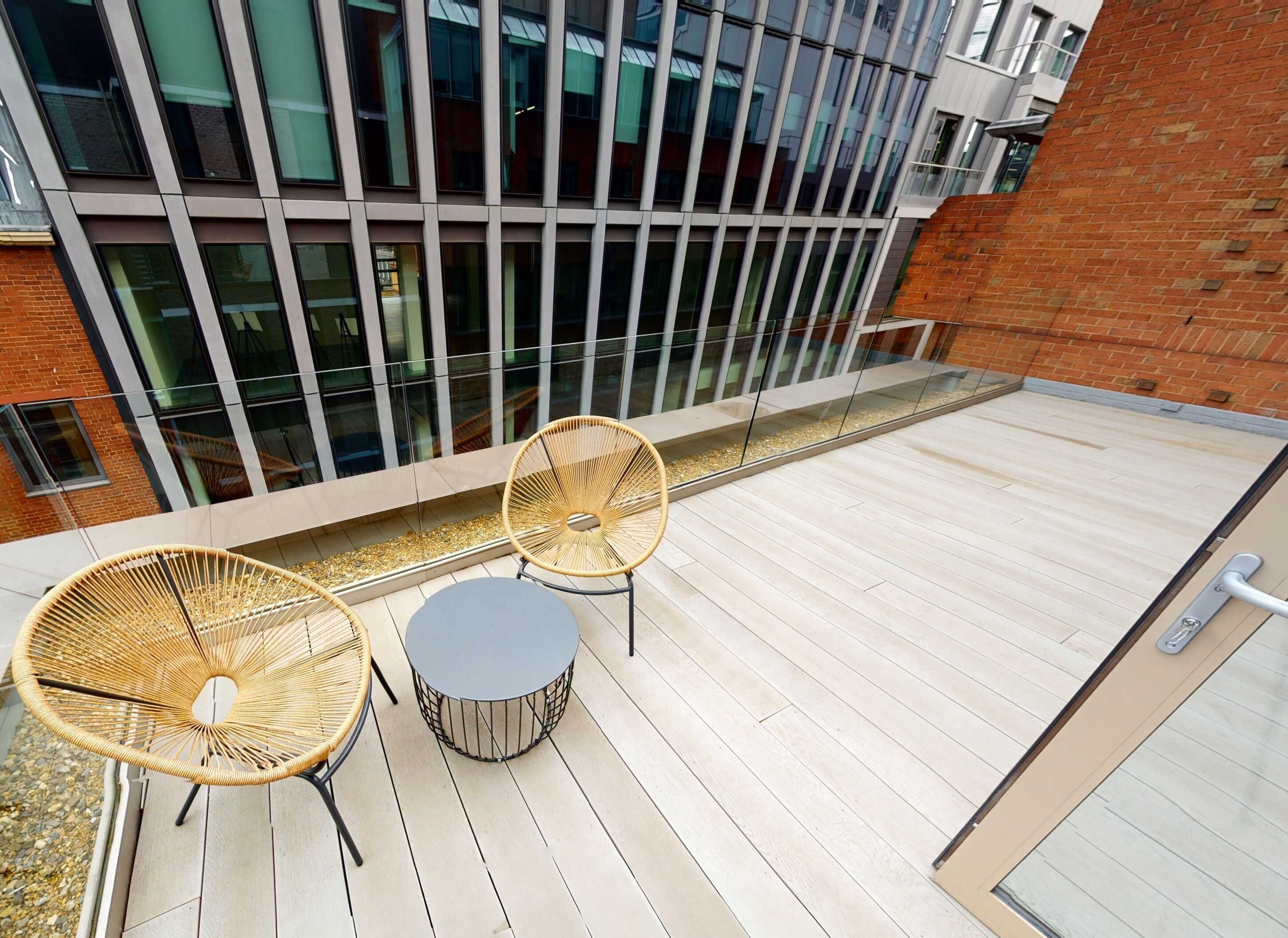 An outdoor terrace with two wicker chairs and a small round table, a glass railing, and a view of a modern building with large windows.