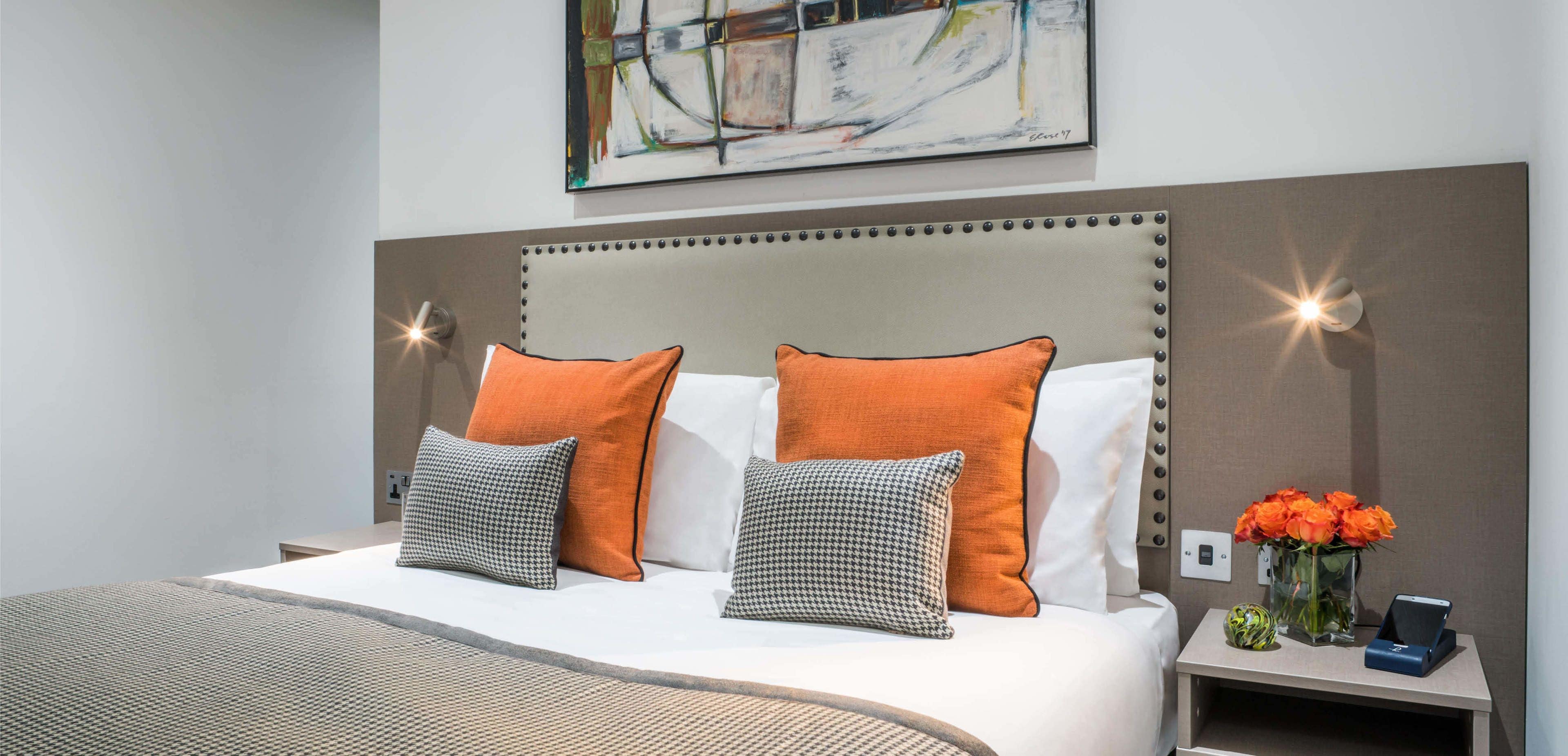 Ein ordentlich bezogenes Bett mit zwei orangefarbenen und zwei grauen Kissen, ein Nachttisch mit einer Vase mit orangefarbenen Blumen und ein an der Wand hängendes Kunstwerk darüber.