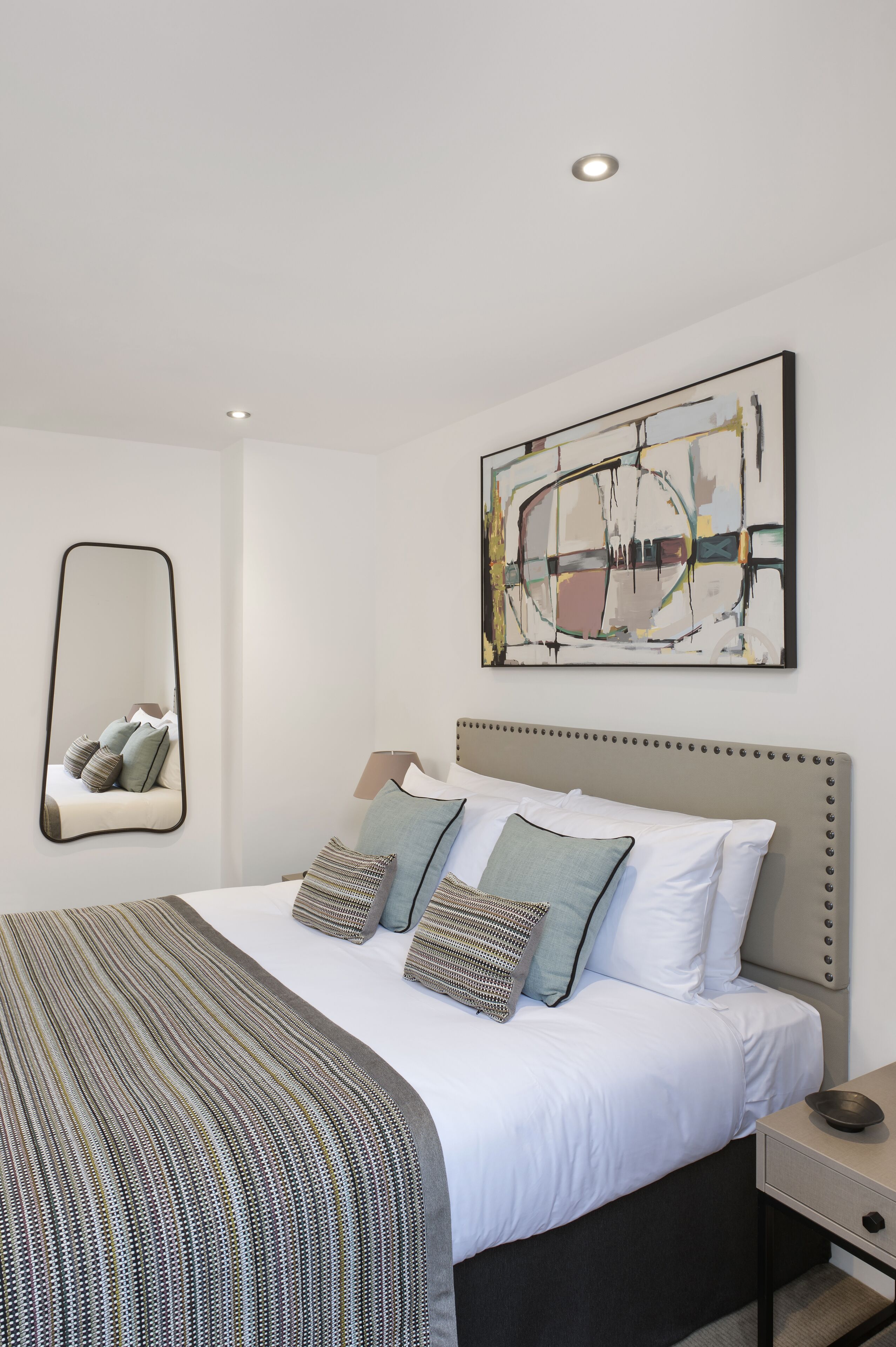 A modern bedroom with a large bed, a bedside table, a mirror, and an abstract painting on the wall.