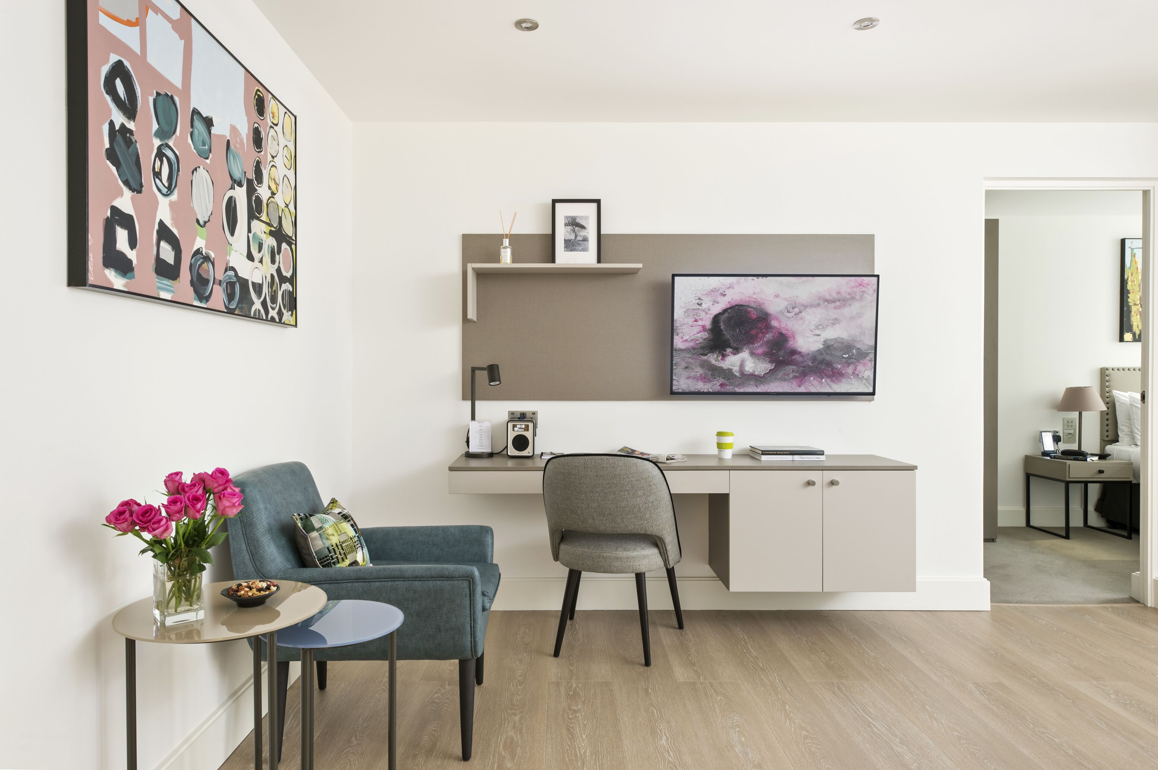 Uma sala de estar moderna com um sofá cinzento, uma poltrona azul, uma mesa redonda pequena com um vaso de flores e uma grande TV de tela plana fixada na parede.