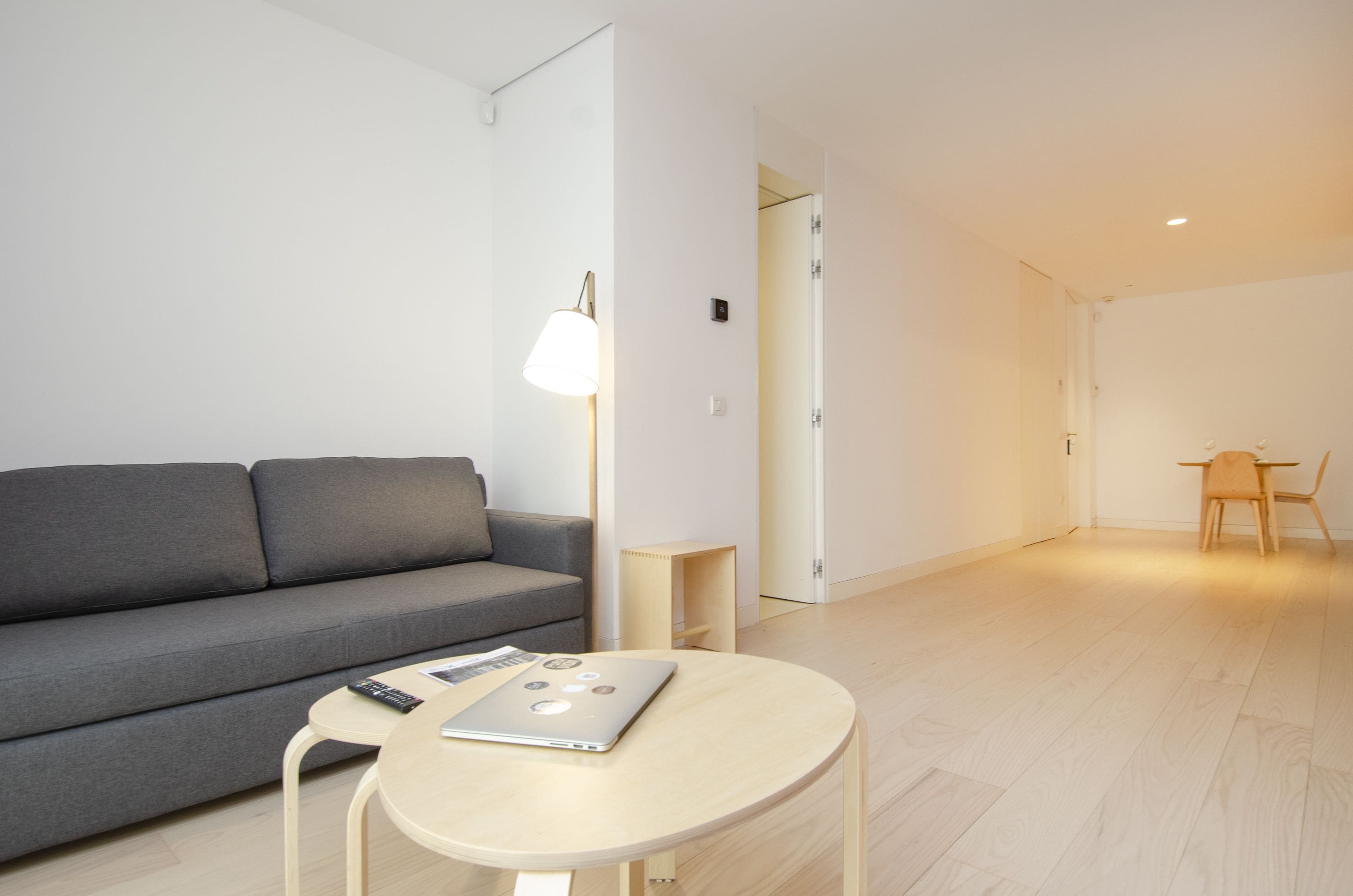 A modern living room with a grey sofa, a round wooden coffee table with a laptop, a small wooden side table, and a lamp.