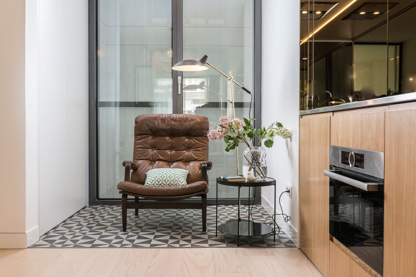 A modern kitchen with a built-in oven, a dining area with a chair and a small table, and a large window with a view.