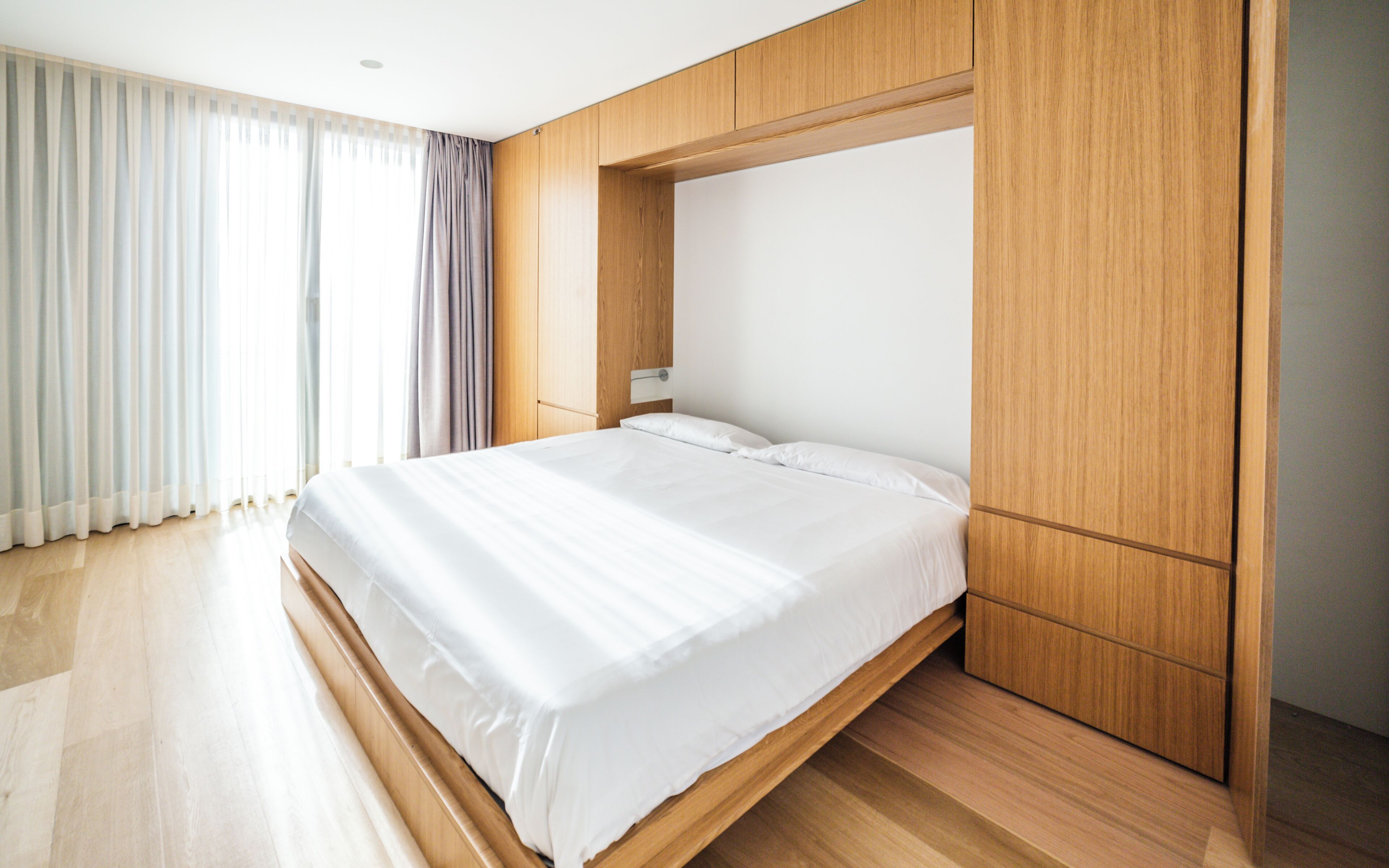 Un dormitorio moderno con un marco de cama de madera, un ventanal con cortinas translúcidas y un armario de madera.