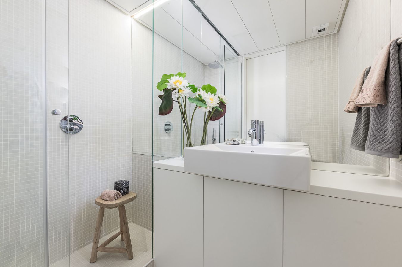 A modern bathroom with a glass shower enclosure, a wooden stool, a sink with a mirror, and a towel rack.