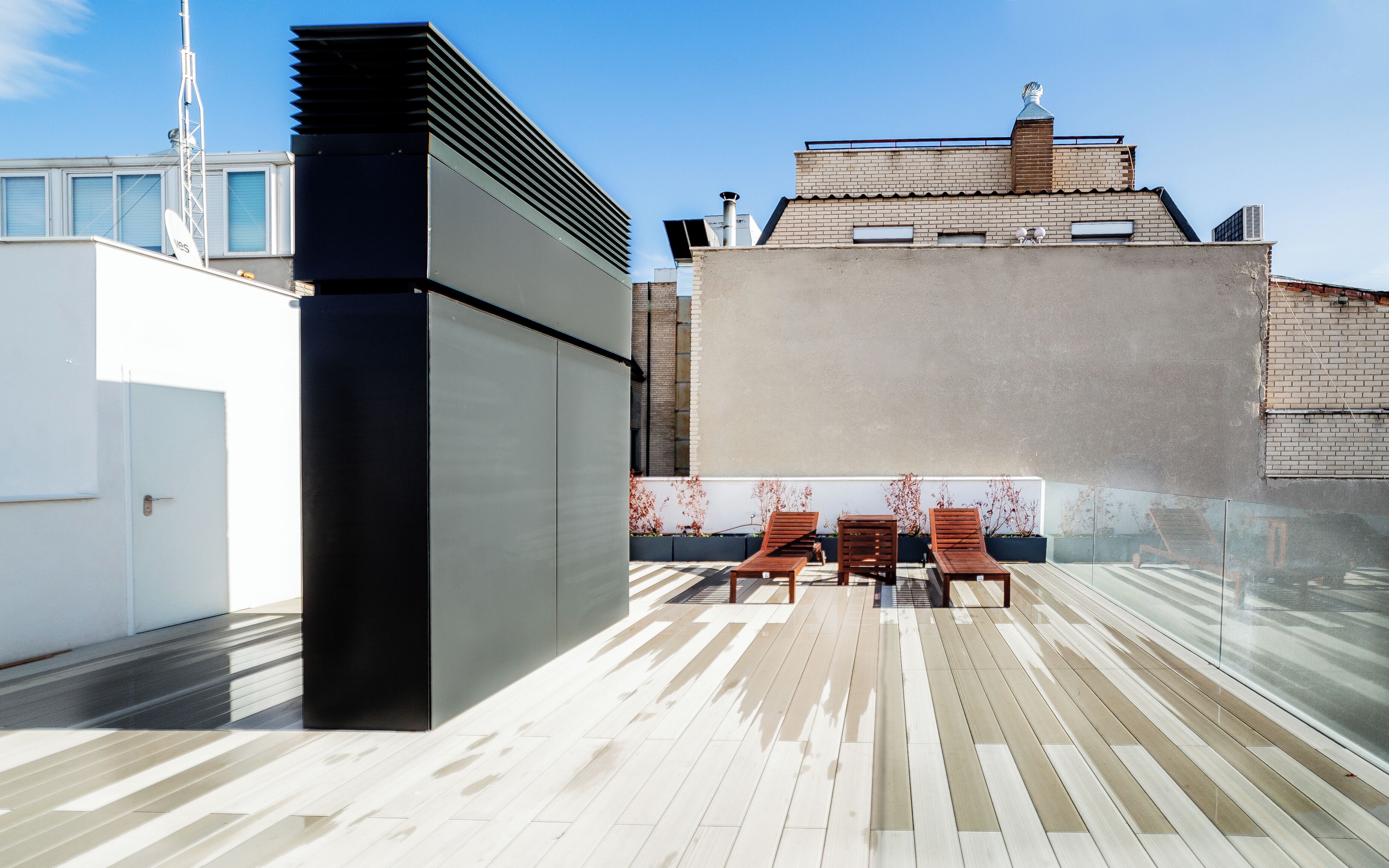 A rooftop terrace with a modern design, featuring a black structure, wooden lounge chairs, and a view of the cityscape.