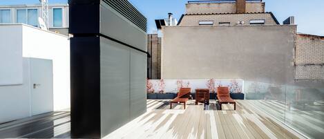 A rooftop terrace with a modern design, featuring a black structure, wooden lounge chairs, and a view of the cityscape.
