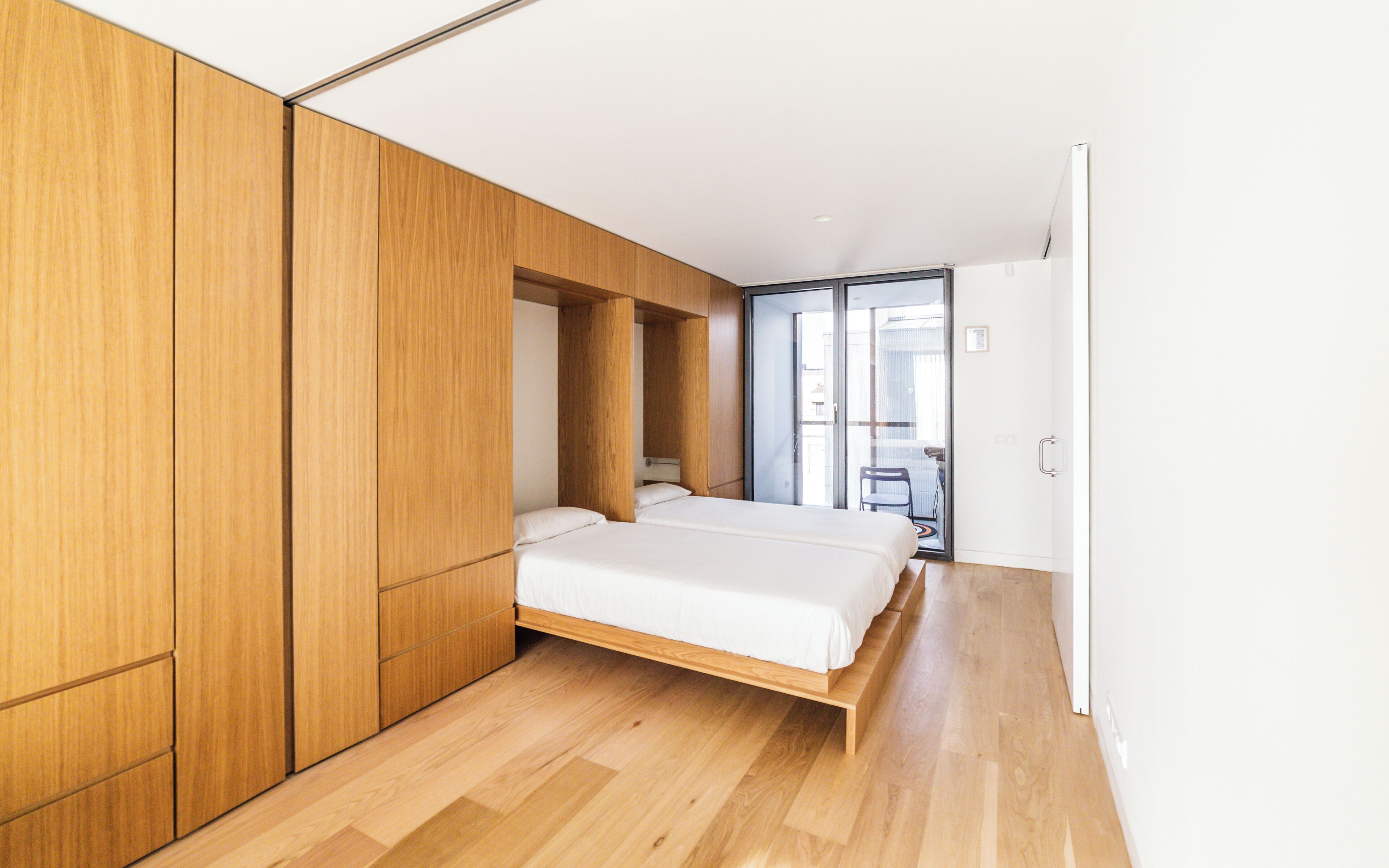 A modern bedroom with a bed, wooden wardrobe, and a sliding glass door leading to a balcony.