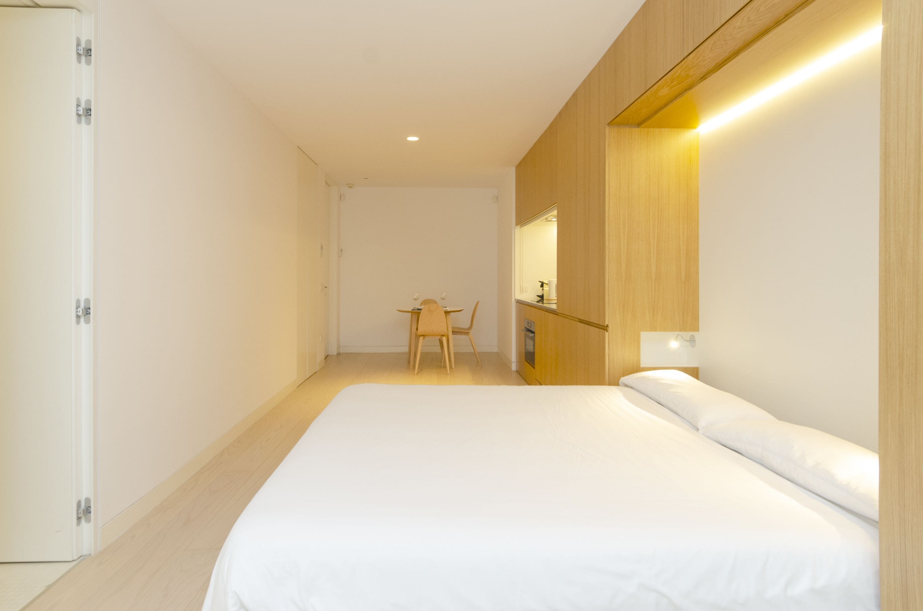 A hotel room with a large bed, a wooden wardrobe, a small desk with a chair, and a mirror above the mirror cabinet.
