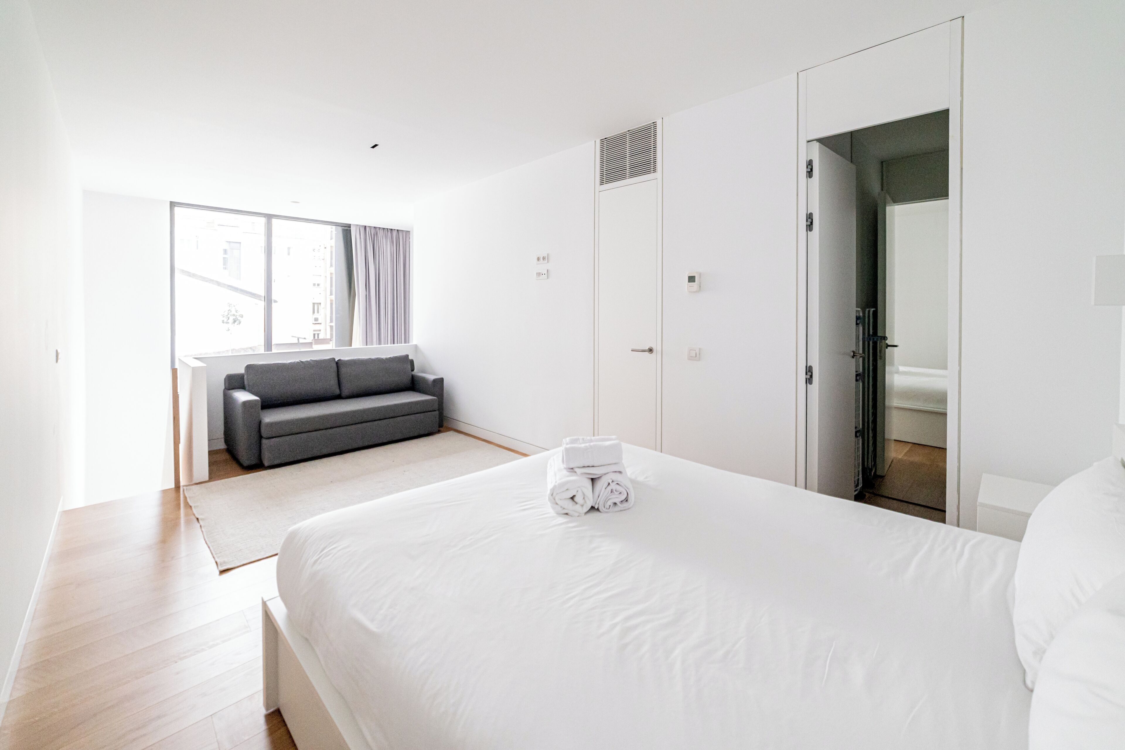 A modern hotel room with a large bed, a grey sofa, and a window with curtains.